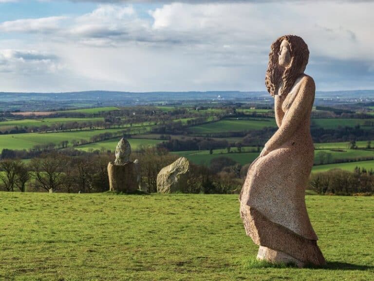 Dans la Vallée des Saints, une sculpture en pierre représentant une personne se dresse gracieusement au milieu d'un champ herbeux, encadré par des collines ondulantes et des rochers dispersés dans un arrière-plan serein.