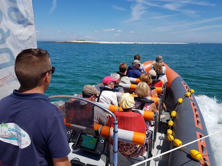 A group of people sit in a dinghy on the blue waters of the ocean, with a guide at the helm, introducing Our Partnerships. The distant shoreline and a few sailboats are visible in the background under a clear sky.