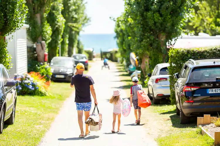 A man and two children walk along a sunny path towards the beach in South Finistère, carrying beach equipment. Cars are parked along the grassy road and trees line the sides, reminiscent of a serene camping getaway.