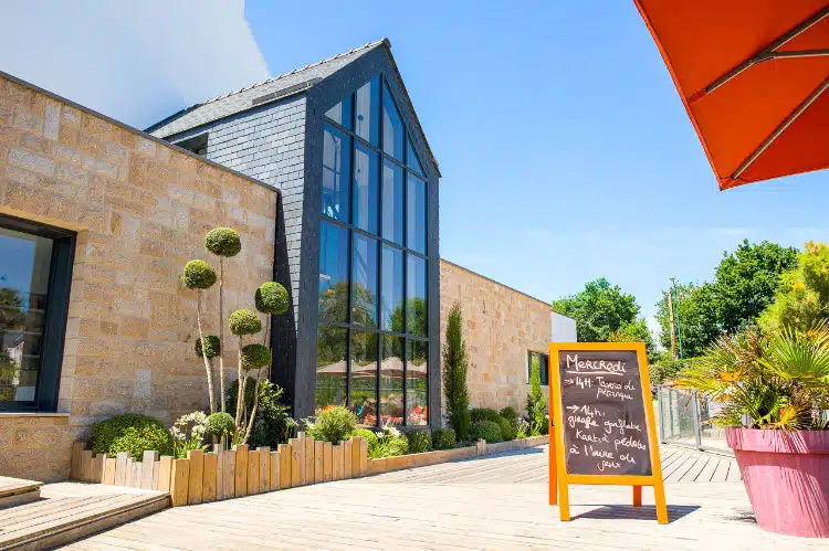 Terrasse de café en plein air avec façade vitrée et mur de pierre rustique, nichée comme un joyau caché rappelant le Finistère Sud. Un menu à l'ardoise et des buissons ronds soigneusement taillés ajoutent du charme, évoquant une ambiance de camping accueillante au cœur de la ville.