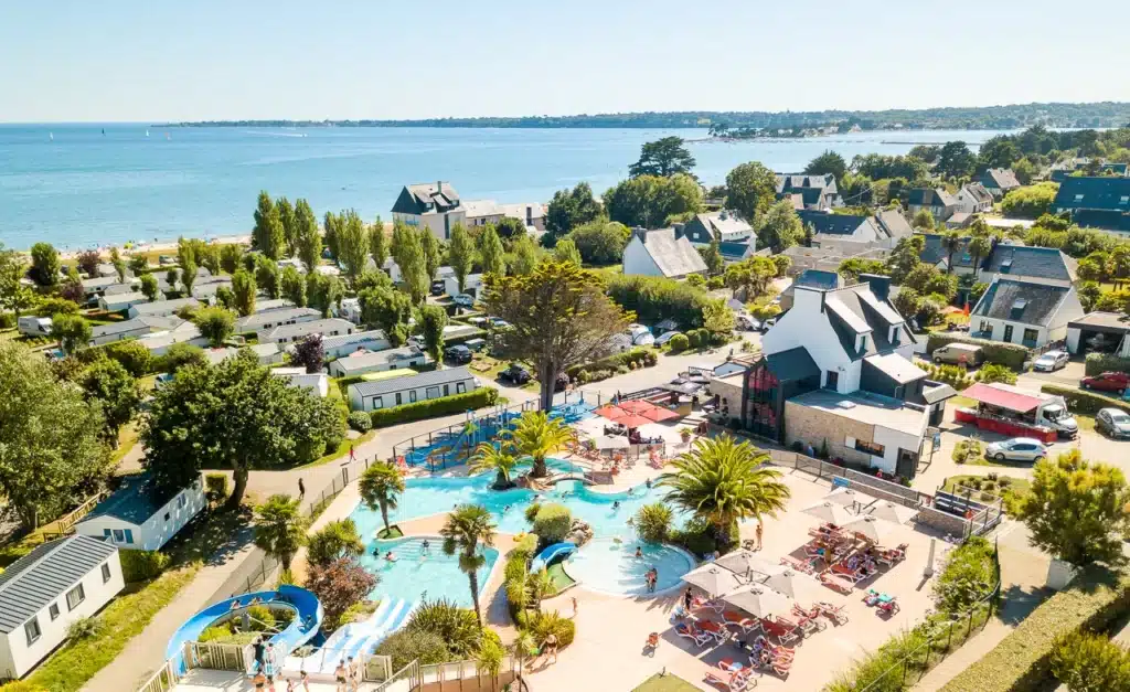 Vue aérienne du camping les Saules, comprenant une piscine, des toboggans aquatiques, des palmiers, des chaises longues et des bâtiments résidentiels environnants en bord de mer.