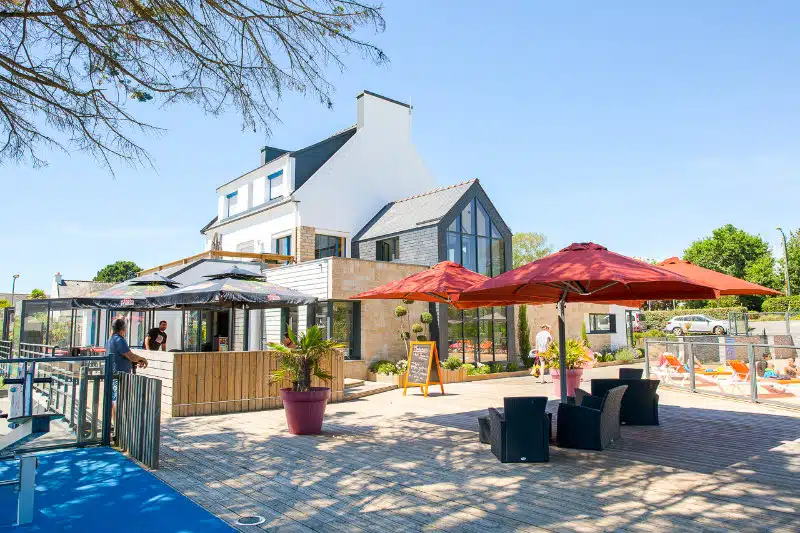 Un bâtiment moderne de deux étages avec des sièges extérieurs sous de grands parasols rouges offre un endroit parfait près de la côte bretonne. Les gens profitent du temps ensoleillé sur la grande terrasse en bois, capturant l'essence de la détente en bord de mer.