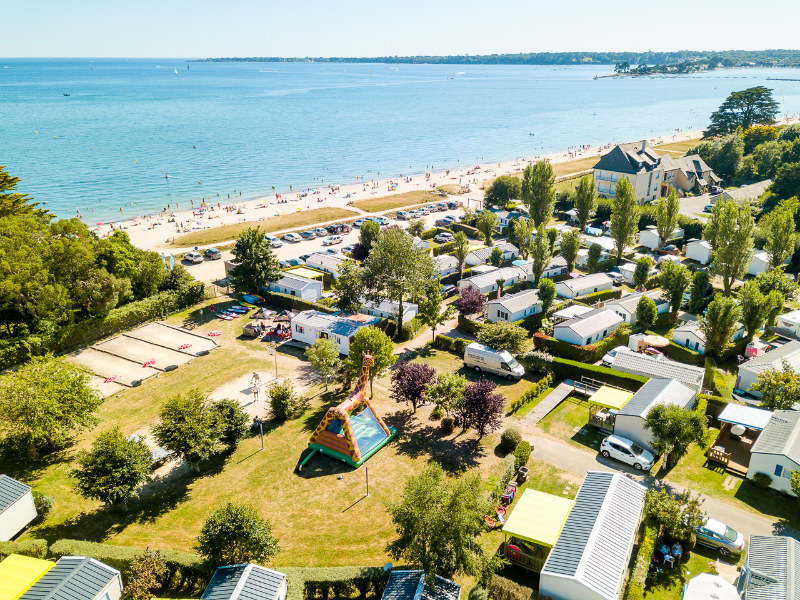 Luftaufnahme eines Campingplatzes am Meer im Finistère mit vielen Wohnmobilen, Bäumen und einem Spielplatz. Im Hintergrund genießen die Menschen den Sandstrand und das ruhige Meer.