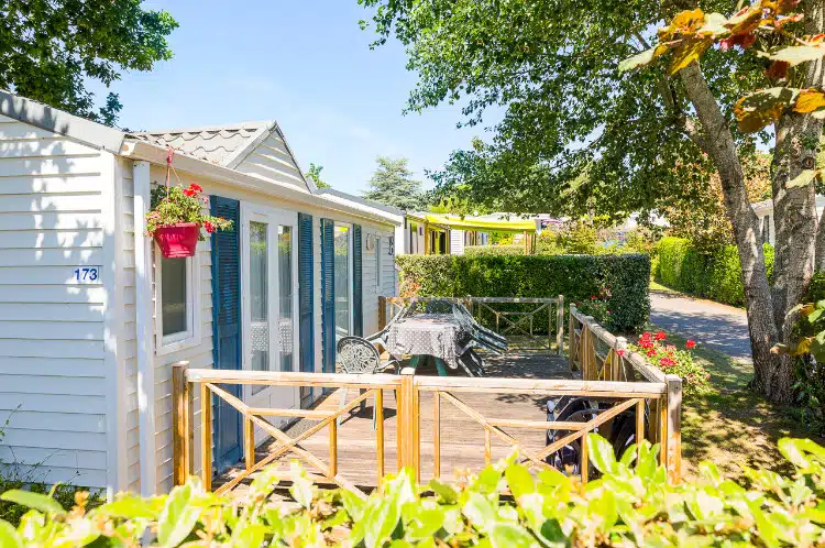 A charming mobile home with wooden terrace, garden furniture and flowerpot, nestled among trees and plants on a sunny day in the heart of Finistère, perfect for a camping holiday.