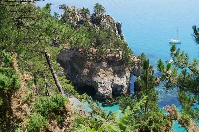 At Cap de la Chèvre, a rocky, tree-lined cliff overlooks clear, turquoise water. Nearby, a sailboat sits peacefully at anchor.