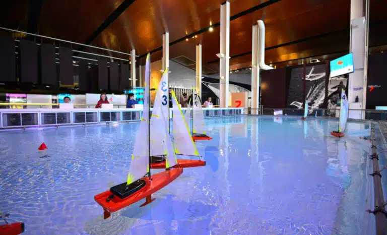 Model sailboats with numbered sails, a tribute to Nos Partenariats, float gracefully on a large indoor pool under a high ceiling enhanced by modern lighting.