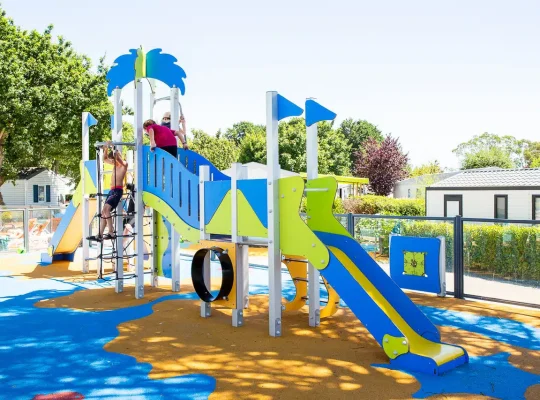 Des enfants jouent sur une structure de jeux colorée avec toboggans et éléments d'escalade, entourés d'arbres et de bâtiments sous un ciel clair, rappelant les voyages de camping en famille dans les paysages sereins du Finistère Sud.