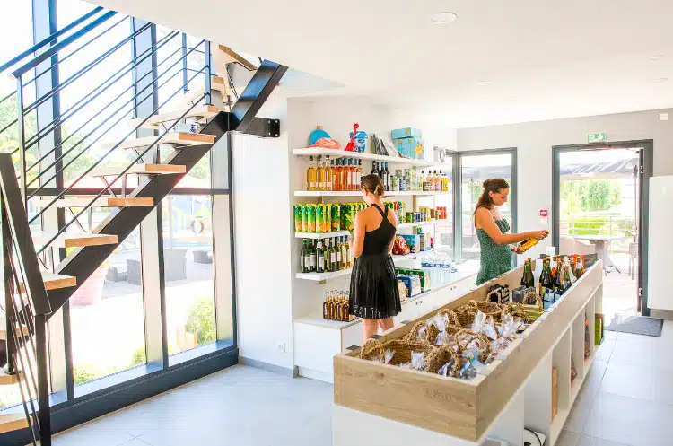 Deux personnes parcourent un magasin lumineux et moderne avec divers produits sur des étagères blanches, parfait pour ceux qui planifient un séjour en camping dans la belle région du Finistère. Un escalier intérieur sur la gauche ajoute au design épuré.