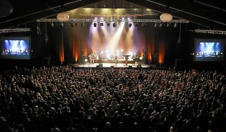 A large crowd gathers for the concert in a dimly lit indoor venue, reminiscent of the Cornouaille Festival, with a brilliantly lit stage and visible performers. Two screens on either side broadcast the performance, capturing the vibrant essence of Brittany at every moment.