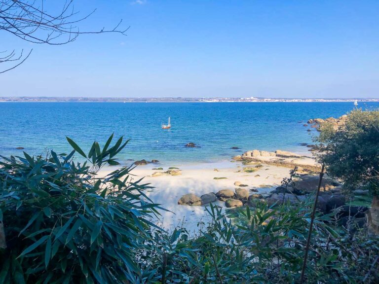 Discover the serene beauty of Beg Meil, where a sandy beach is framed by lush vegetation and rocks. A small sailboat glides on the calm blue sea under a clear sky, offering perfect conditions for leisure activities. The picturesque coastline beckons you away, inviting exploration and sightseeing.