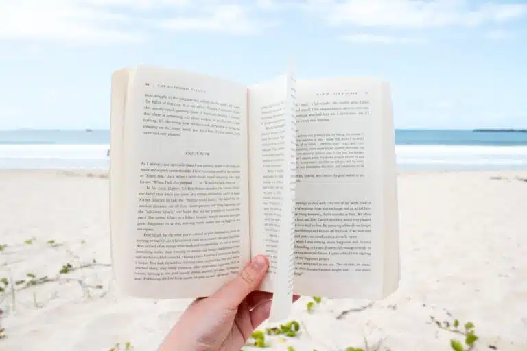 Une personne profite de ses vacances, tenant un livre ouvert sur une plage de sable avec l'océan et un ciel bleu nuageux en arrière-plan, perdue dans un monde de feu.