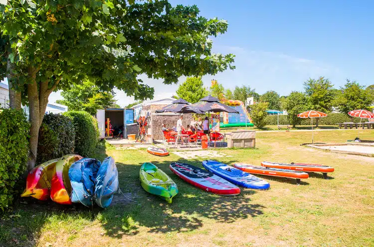 Kayaks and paddleboards rest on the grass next to a shaded lounge area with tables and umbrellas, perfect for a camping retreat. Surrounded by trees under the clear blue skies of South Finistère, it's an idyllic getaway.