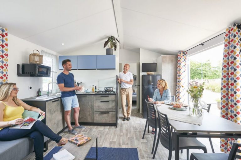 In a modern kitchen and dining room reminiscent of a mobile home, four people gather. One person stands near the kitchen, two sit at the dining table and another relaxes on the sofa with a magazine.