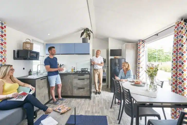 Dans une cuisine et une salle à manger modernes rappelant un mobil-home, quatre personnes se réunissent. Une personne se tient debout près de la cuisine, deux sont assises à la table à manger et une autre se détend sur le canapé avec un magazine.