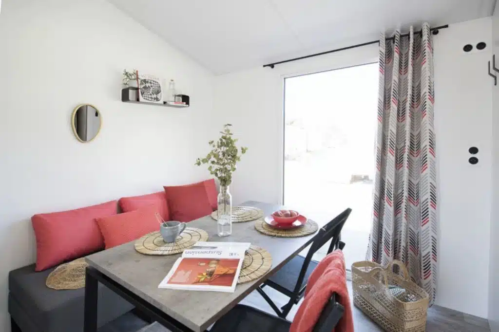 A cozy dining area in a Fouesnant mobile home, with a wooden table set for two, red cushions, a vase of greenery, a magazine and textured curtains framing the large window.