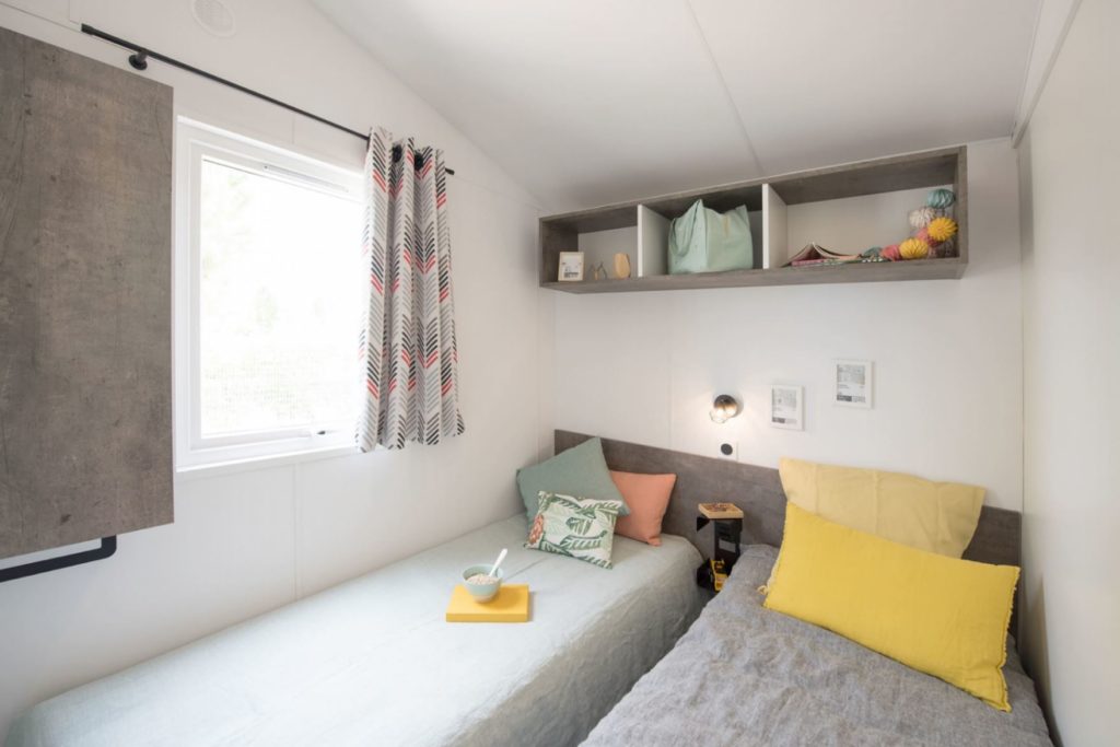 A comfortable room in a mobile home in Fouesnant features two single beds with green and yellow blankets, a window and a bookshelf. A bowl rests on one of the beds, while the shelves display books and decorative objects. A light fixture hangs over the beds.