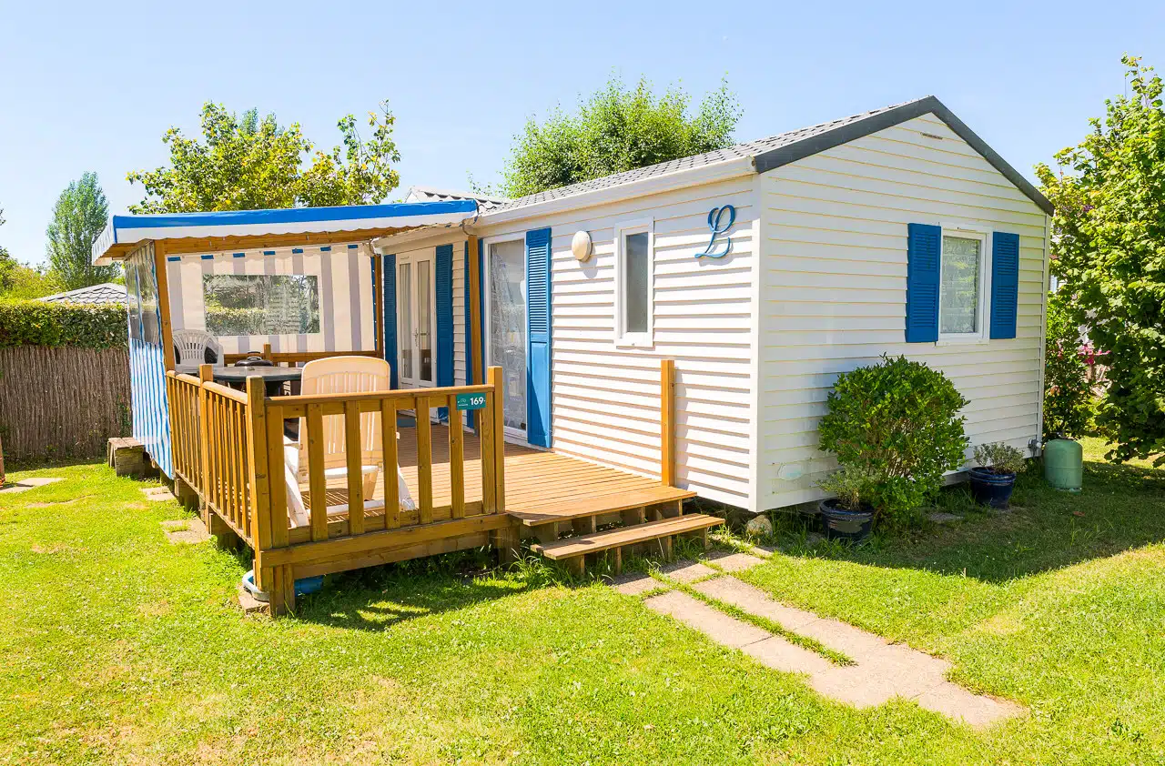 Un petit mobil-home blanc aux volets bleus et terrasse en bois est niché dans un espace herbeux et arboré, reflétant l'essence du camping du Sud Finistère. Une table et des chaises vous invitent à vous détendre sur la terrasse au cœur de la nature.