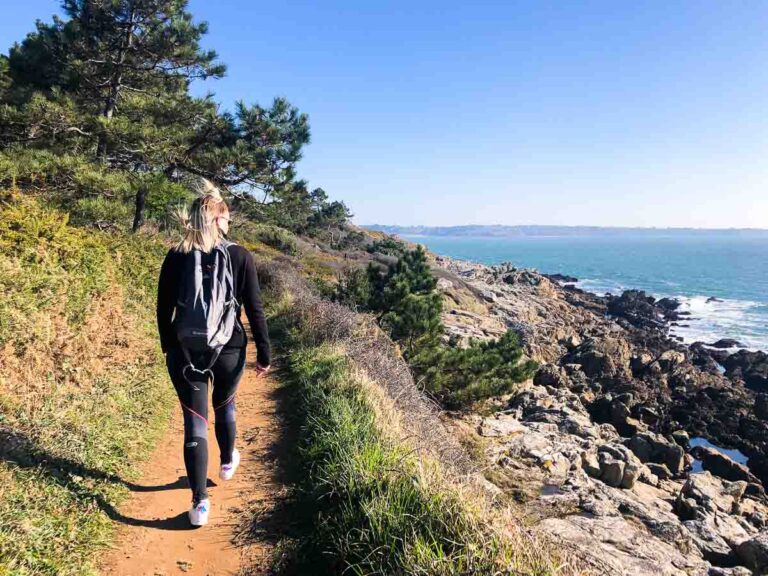 A person with a backpack walks along the Nevez coastal path, traversing rocky terrain and trees with the ocean in the background on a clear day.