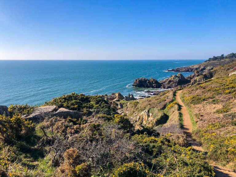 Unter dem klaren blauen Himmel eines typischen Tages in Nevez enthüllt sich die Küstenlandschaft mit einem Feldweg, der sich an zerklüfteten Felsklippen entlang schlängelt, während sich der Ozean bis zum Horizont erstreckt.