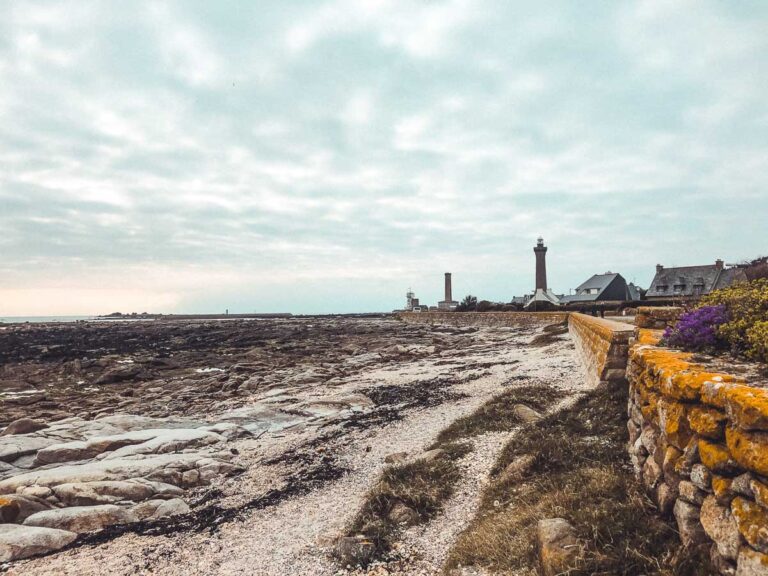 Entdecken Sie Penmarc'h und seine spektakuläre Küstenlandschaft mit den felsigen Küsten und dem imposanten Leuchtturm im Hintergrund. Eine Steinmauer säumt die malerischen Häuser unter einem bewölkten, trüben Himmel und verkörpert den einzigartigen Charme des Tourismus in Penmarc'h.