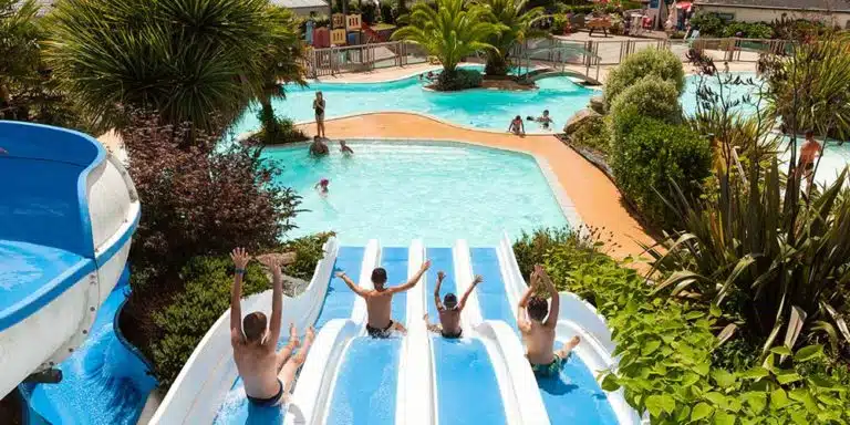 People slide down waterslides in a water park, splashing in a pool surrounded by lush greenery and palm trees, creating the perfect oasis for those camping nearby.