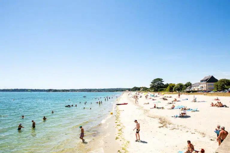 People enjoy a sunny day at the Finistère beach, swimming in the sea and relaxing on the sandy shore, with a few buildings in the background.