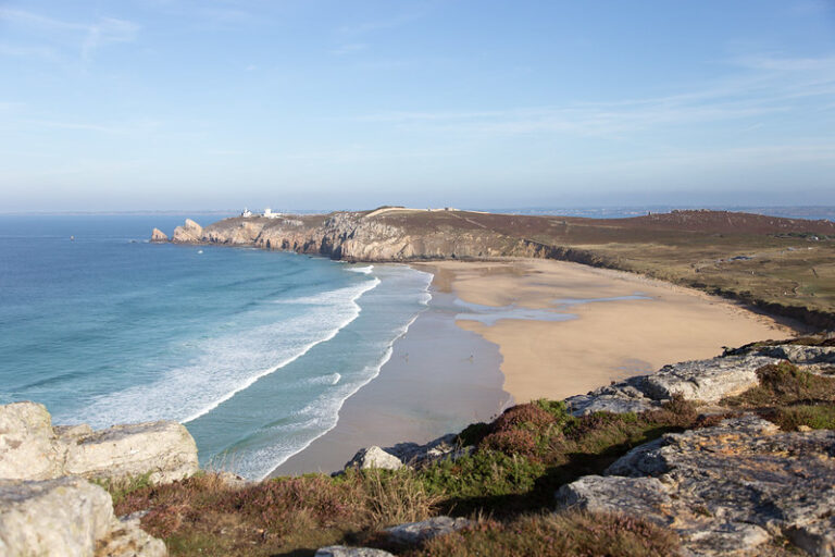 Un paysage côtier pittoresque se dévoile à La Pointe de Pen Hir, où une plage de sable rencontre des falaises majestueuses et des vagues dansent sous un ciel bleu clair.