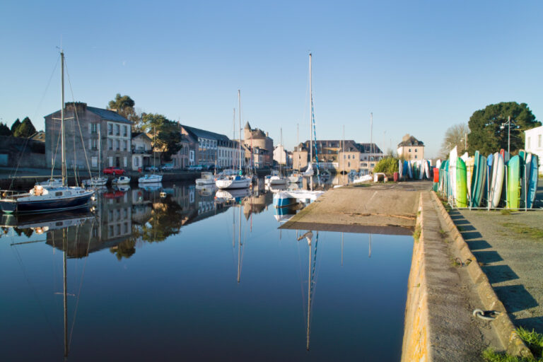 Une scène de port paisible avec des voiliers amarrés, entourés de vieux bâtiments en pierre sous un ciel bleu clair. Une rangée de planches de surf se dresse sur la droite, capturant l'essence d'un carnet de voyage à travers le charmant Pays Bigouden.