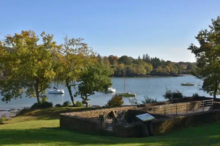 Un pont de pierre serein s'étend gracieusement sur l'herbe luxuriante près d'une rivière bordée d'arbres, où plusieurs voiliers glissent paisiblement. Ce cadre idyllique reflète presque les paysages souvent capturés dans les œuvres de Romain Perrenou.
