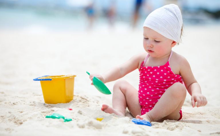 A toddler in a pink polka-dot bathing suit and white hat plays with a yellow bucket and green shovel on a sandy beach, capturing the joyful essence of baby life in Southern Brittany.